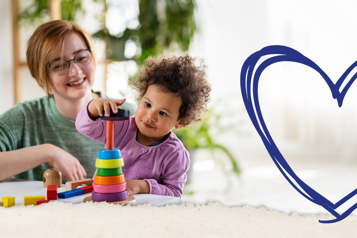 A caregiver smiles while watching a young child stack colourful rings on a toy, with a large blue illustrated heart on the right side of the image.