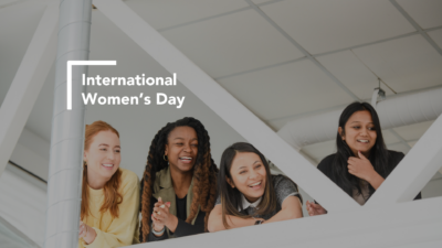 Text on the graphic says "International Women's Day" and picture in the background is of four women leaning over a railing watching something below and smiling.