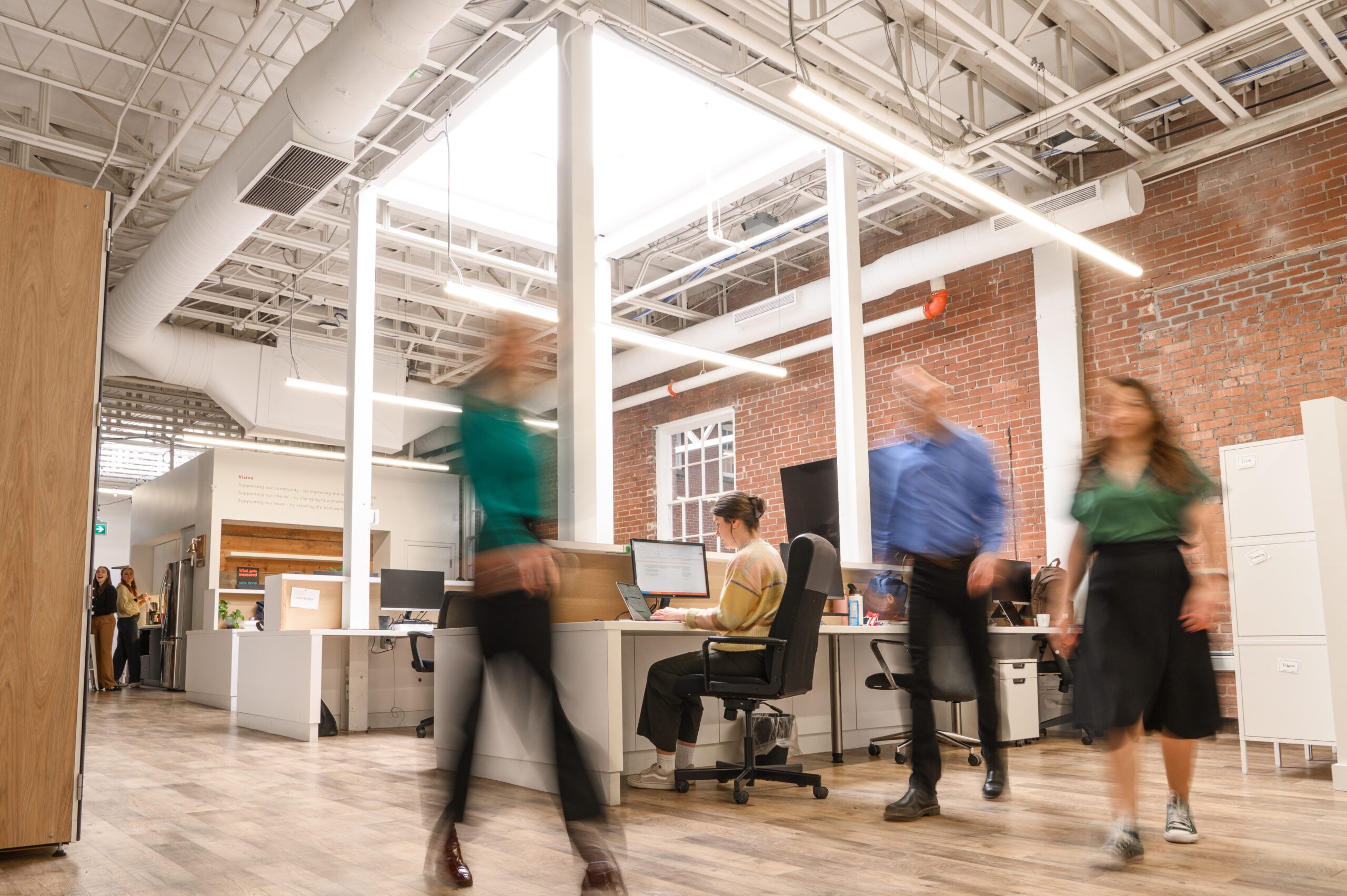 A photo of a busy office environment. People are moving through the space in all directions and they are blurred because they are in motion
