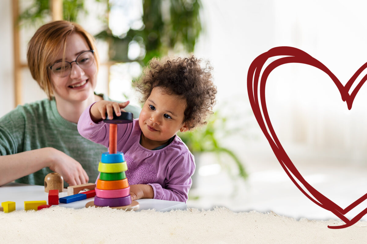 Image shows a woman watching a child play with a stacking toy. There is a heart graphic in the right hand corner.