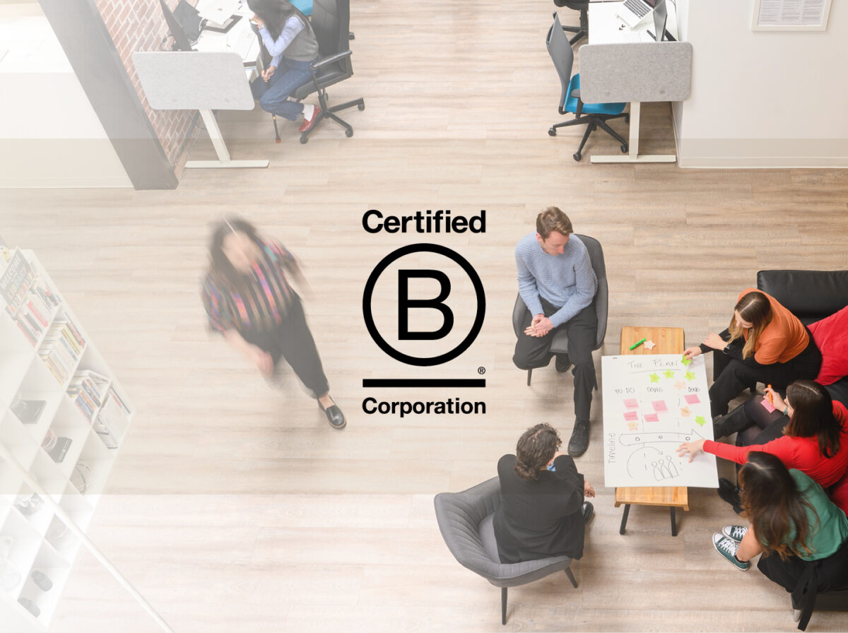 An overhead shot of a group of employees meeting to do an engagement session while Emily walks by to another meeting. The B Corp Logo is imposed on the left side.