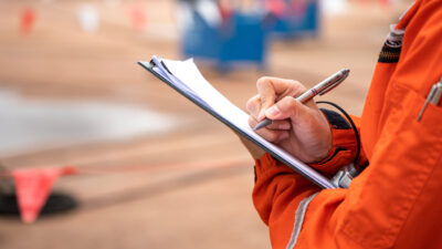 A close up of an inspector. They are wearing an orange safety vest and holding a clipboard
