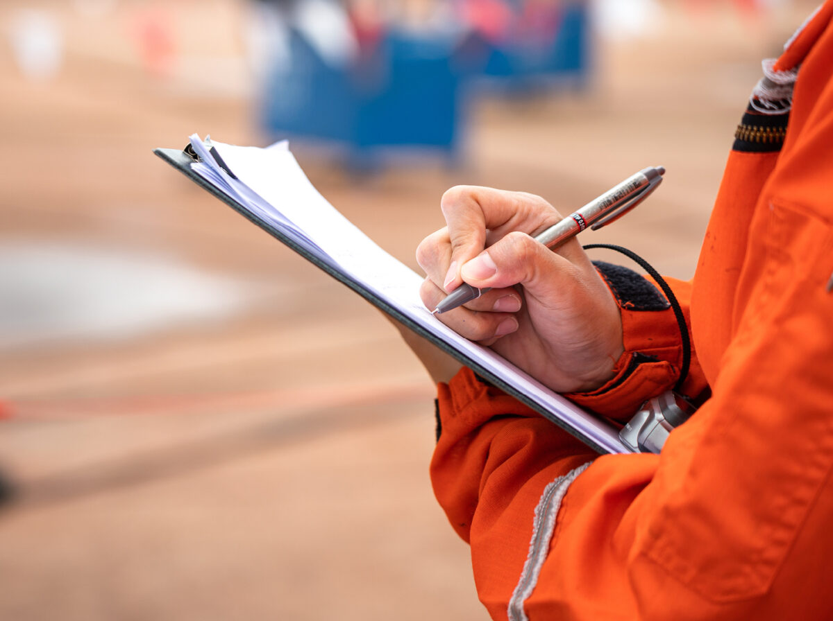 A photo of a inspector on a worksite. It is a close up photo of a person in an orange jacket holding a clipboard and a pen
